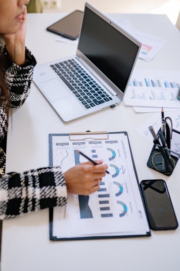 Person analyzing business data on paper with charts next to a laptop in an office setting.